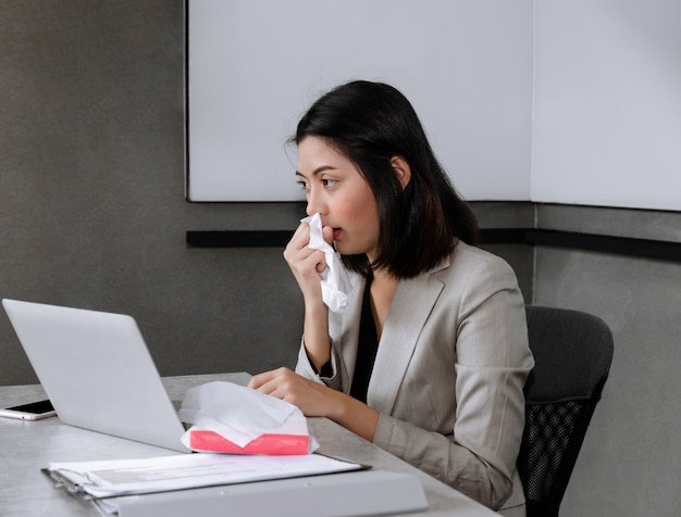 Sick businesswoman sitting at office desk with laptop, sneezing and coughing