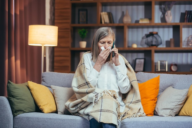 Sick grayhaired woman at home has a runny nose sits on the couch calls the doctor for help and advice