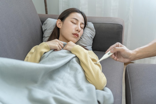 Sick influenza asian young woman girl headache have a fever flu and check thermometer measure body temperature feel illness sitting on sofa bed at home Health care person on virus covid19