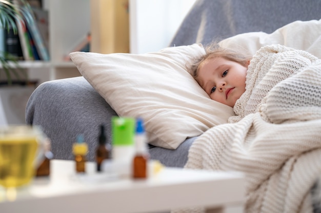 Sick little girl is lying on sofa, wrapped in a blanket. on the table medicines.