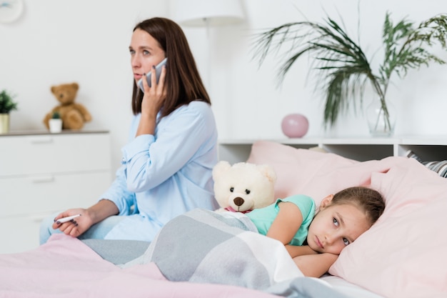 Sick little girl lying on bed under blanket while her mother calling doctor on mobile phone