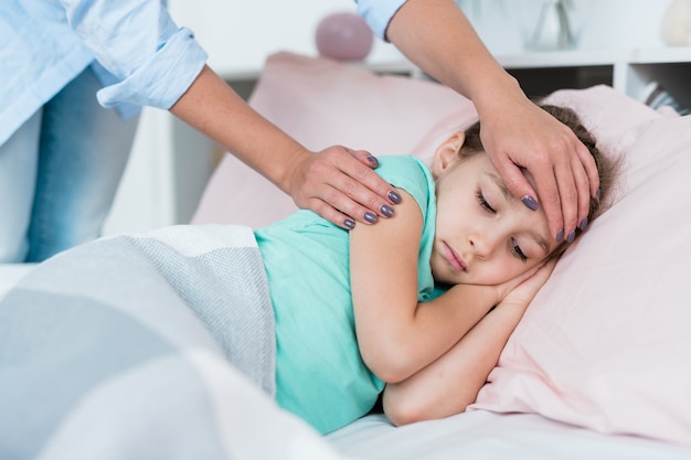 Sick little girl lying in bed under blanket while her mother touching forehead to see if the temperature in high