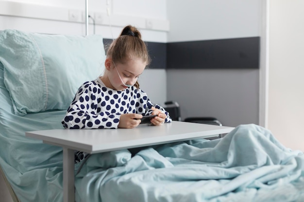 Photo sick little girl playing on smartphone while resting on patient bed in pediatric clinic room. ill young patient enjoying mobile phone entertainment while sitting in hospital recovery ward.