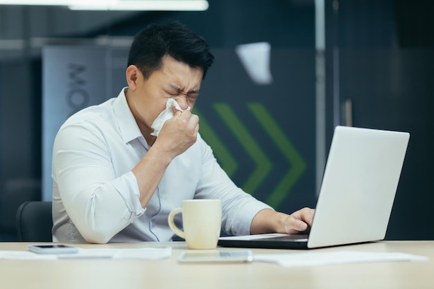 Sick man in office coughs and has runny nose asian businessman with cold works in office on laptop