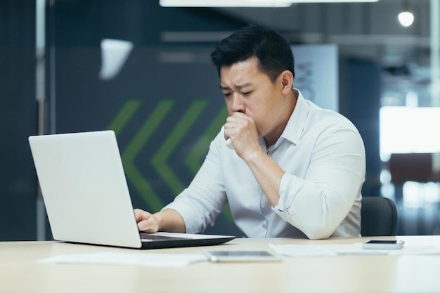 Sick man in office coughs and has runny nose asian businessman with cold works in office on laptop