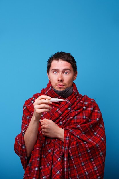 A sick man wrapped in a blanket is standing with a thermometer in his hands