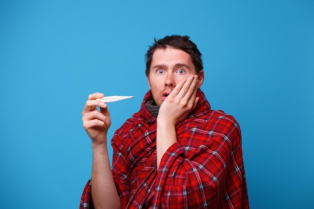 A sick man wrapped in a blanket is standing with a thermometer in his hands