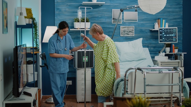 Sick old woman walking with iv drip bag and nurse giving assistance