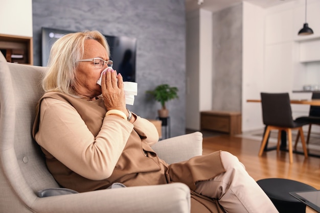 Sick senior woman sitting in chair at home and blowing her nose.