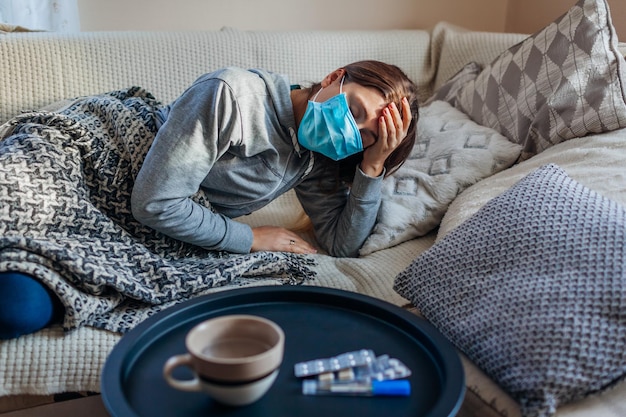 Sick woman having flu or cold Girl lying in bed wearing protective mask by pills and water on table