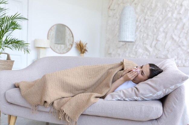 A sick woman is lying under a blanket on the sofa at home alone has a cold is sneezing