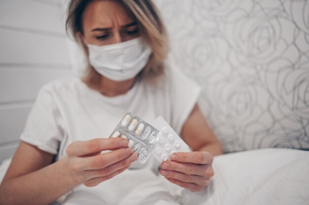 Sick woman lying in bed holding pills at home quarantine isolation. Corona virus COVID-19 concept. Infection causes respiratory illness first symptoms