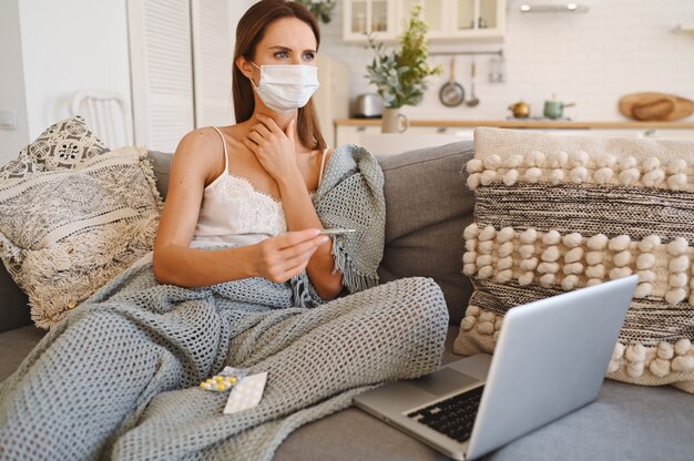 Sick woman wearing face protection mask, sitting on couch with laptop and holding thermometer and pills during home quarantine self isolation. 