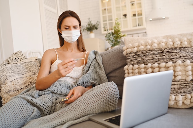 Sick woman wearing face protection mask, sitting on couch with laptop and holding thermometer and pills during home quarantine self isolation. 