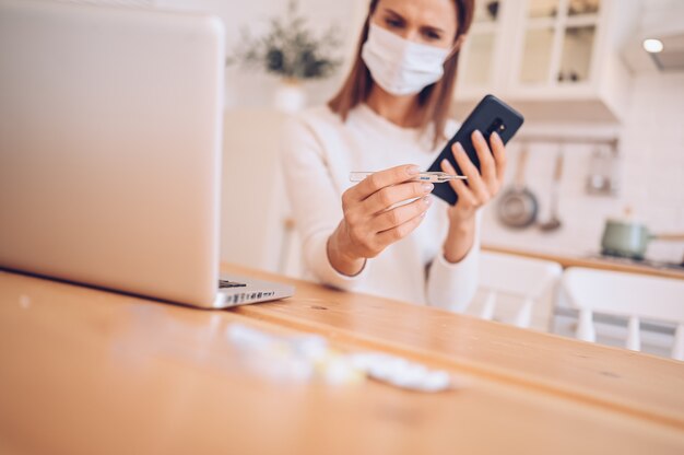 sick woman wearing medical mask, speaking smartphone and working on a laptop in kitchen home during quarantine isolation Covid-19 pandemic 