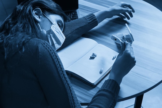 Sick woman with pills and thermometer on a table working on a laptop from home on quarantine