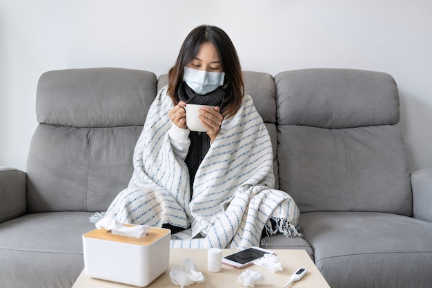 Sick young Asian girl having influenza symptoms wearing medical mask while hold a cup of hot water sitting on sofa with tissue, medicine and thermometer on desk. Cold, flu and home isolation concept.