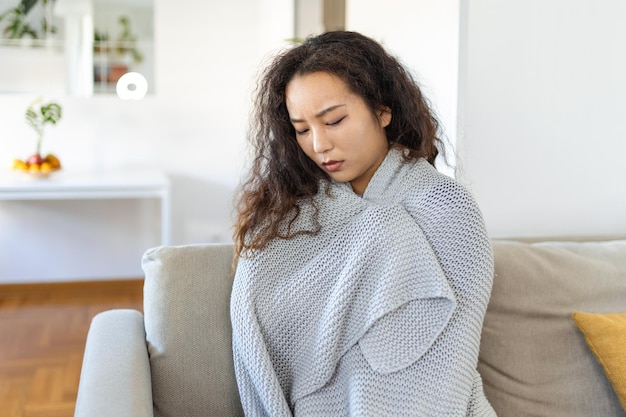 Sick young Asian woman feeling cold covered with blanket sit on bed ill black girl shivering freezing warming at home wrapped with plaid no central heating problem fever temperature flu concept