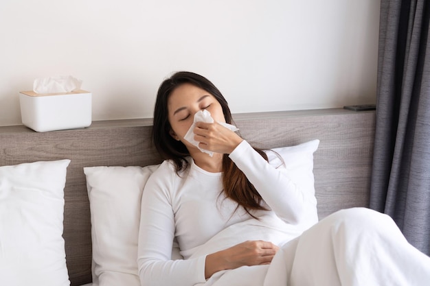 Sick young Asian woman feeling cold and has a high fever blowing nose while lying down on bed in the morning at home Allergy flu concept