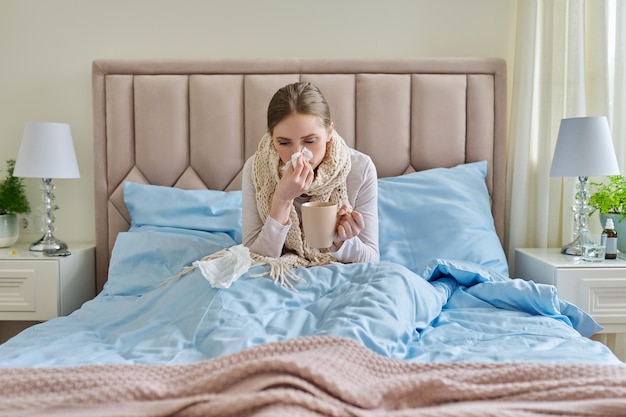 Sick young woman at home in bed with cup of hot drink and handkerchief