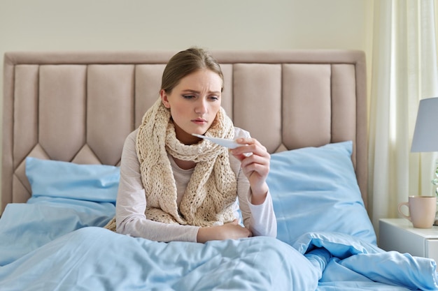 Sick young woman with thermometer in her hands sitting at home in bed