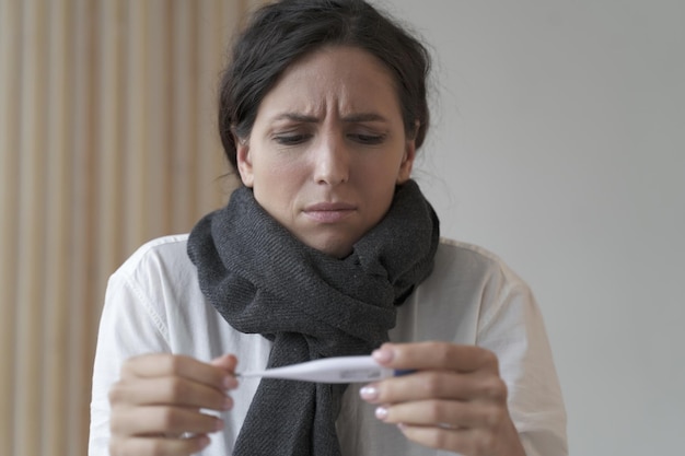 Sicklooking young business woman in knitted scarf looks frowning at electronic thermometer