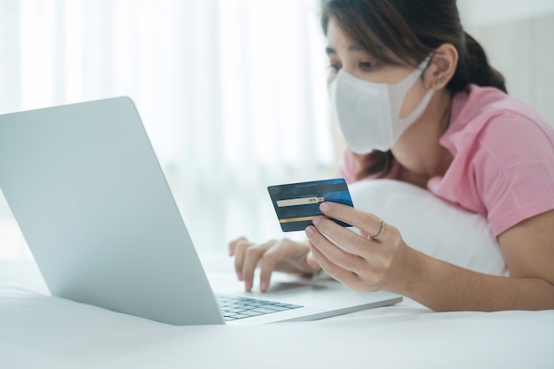 Sickness woman with face mask holding credit card and using computer laptop for online shopping on bed in morning at home. 