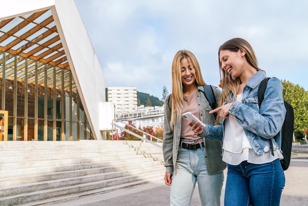 Side front view of a young woman using her mobile phone and pointing at something on the screen showing something funny to her companion walking with her on campus with copy space.