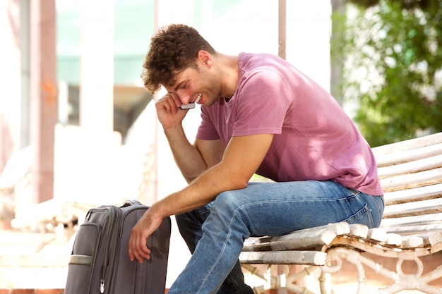 Side of laughing man sitting on park bench with luggage and cellphone