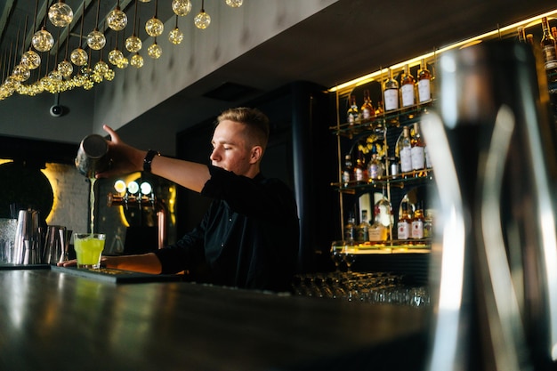 Side lowangle view of barman pouring fresh alcoholic drink into glasses with ice cubes behind bar counter in modern nightclub