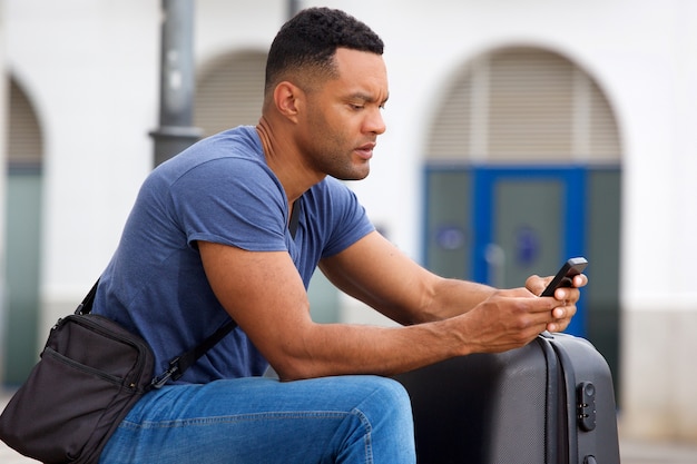 Photo side portrait of african man sitting outside with suitcase and using cell phone