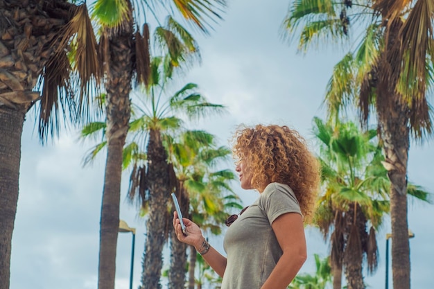 Side portrait of tourist using mobile phone in outdoor with palms street road in backgorund One female people messaging happy Concept of tourism and summer tropical holiday vacation leisure