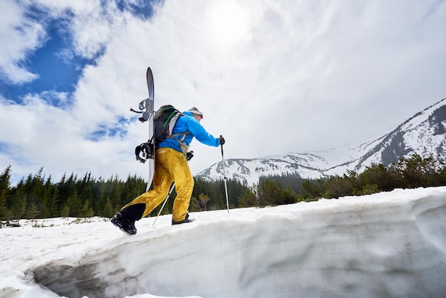 Side of snowboarder hidden face with snowboard on his back on snowy slope Clouds on blue sky on background Low angle