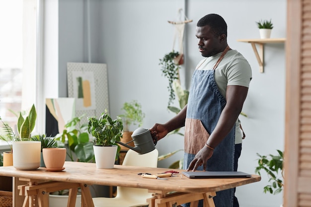 Side view adult black man watering plants indoors and wearing apron