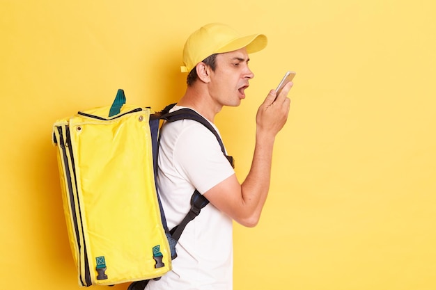 Side view of aggressive courier man with thermo backpack on his back delivering fast food holding smart phone and screaming with anger talking to client or boss isolated over yellow background