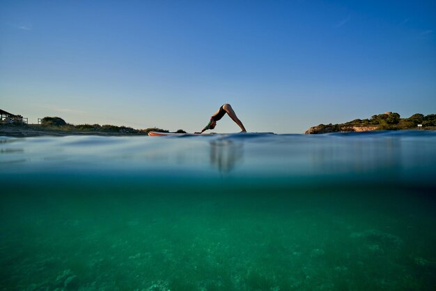 Side view of anonymous female athlete in swimwear practicing yoga on SUP board and taking Adho Mukha Svanasana pose among rippling turquoise ocean