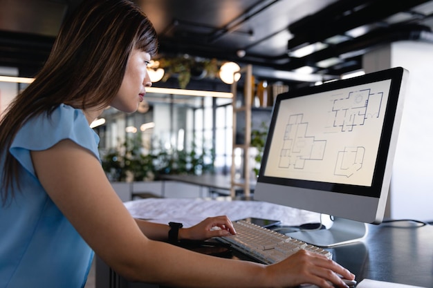 Side view of an Asian businesswoman wearing smart clothes and glasses, working in the modern office, sitting on a desk and using her computer.