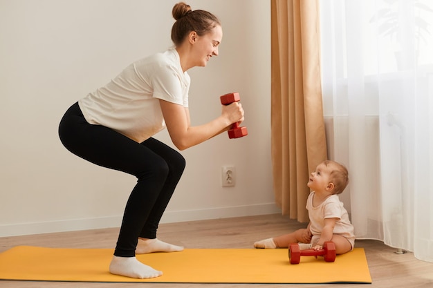 Side view of athletic woman wearing white t shirt and black leggins doing squatting with dumbbell in hands looking with happy expression at baby daughter near her