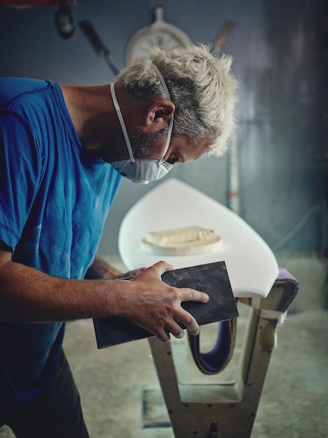 Side view of attentive young male master with blond hair and tattoos on hands in casual clothes and respirator polishing edges of surfboard using sandpaper in workshop