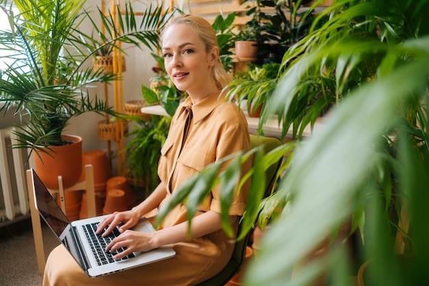 Side view of attractive business woman floral store owner using laptop sitting on background of green plants