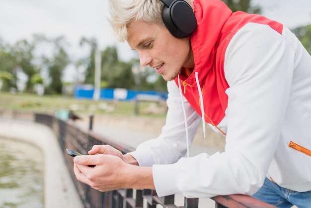 Side view of attractive Caucasian man messaging via social networks at smart phone and using headphones for listening to music in front the lake in the park outdoor People lifestyle concept