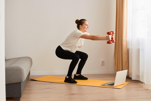 Side view of attractive woman making deep squats with dumbbells in hands in living room while watching fitness online lesson on laptop wearing white t shirt and leggins