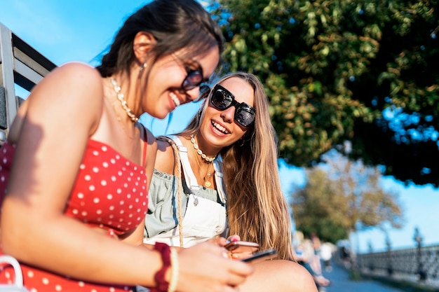 Side view of attractive young female friends using mobile phones while hanging out on city street during summer