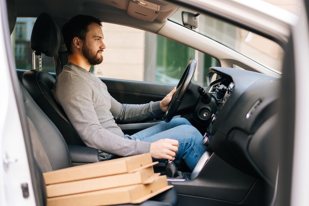 Side view of bearded delivery man wearing casual clothes driving car delivered hot pizza to customer. Concept of fast online delivery around the city.