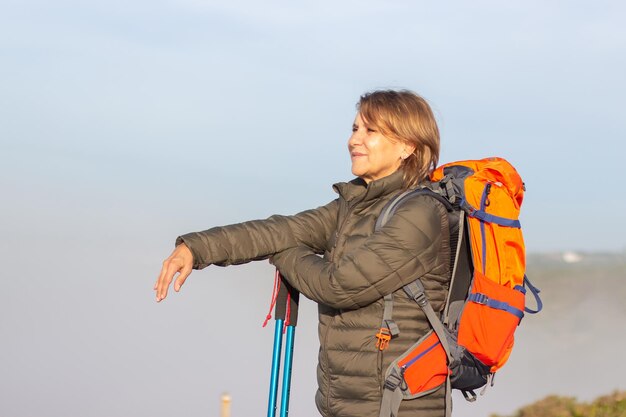 Side view of beautiful female hiker on sunny day. Woman with big orange backpack standing at meadow, looking at landscape. Hobby, active lifestyle concept