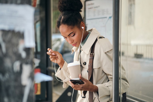 Side view of beautiful girl in stylish trench coat with coffee and cellphone at bus stop