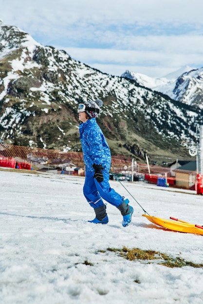 Side view of boy with plastic sledge walking up snowy slope while spending time on sunny winter day in highlands