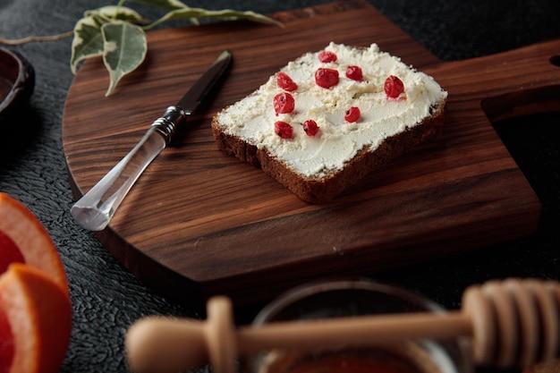 Side view of bread slice smeared with cottage cheese with red currants on it and knife on cutting board on black background