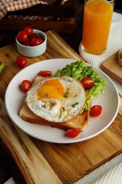 Side view of breakfast set with fried egg lettuce tomatoes on dried bread slice in plate on cutting board with orange juice on cloth and box of bread slices on wooden background