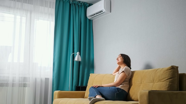 Side view of a brunette woman adjusts the air conditioner while sitting on the sofa